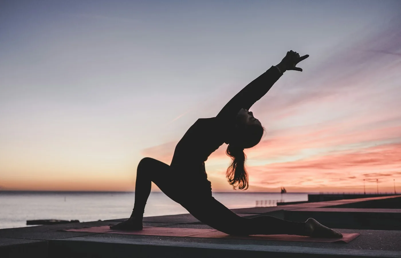 Person übt Yoga auf einer Matte bei Sonnenuntergang am Wasser